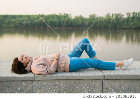 Woman lying down on embankment by lake or river at city park - dreamer and relax concept Woman lying down on embankment by lake or river at city park - dreamer and relax concept 103063915