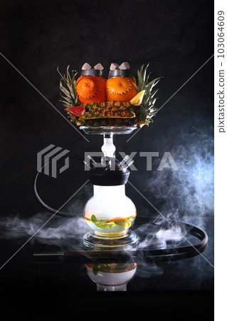 Hookah on pineapples and oranges. A smoking device. Hookah on a black background with a mixture of citrus and pineapple. The concept of smoking and having fun.Smoke on black background. 103064609