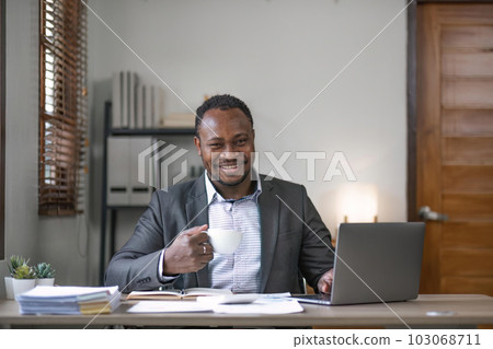 Young African black mansmiling businessman drinking coffee and using laptop in office, sitting at desk 103068711