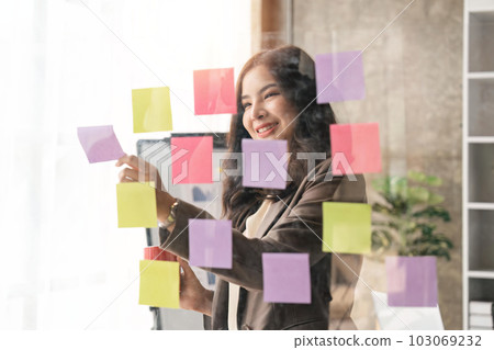 Business Asian woman use post it notes in glass wall to share idea. Brainstorming business office concept. Sticky note on glass wall 103069232