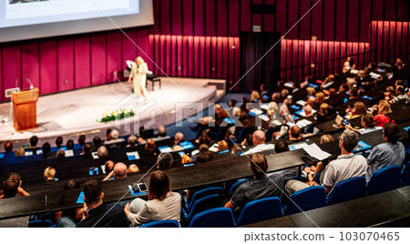 Business and entrepreneurship symposium. Female speaker giving a talk at business meeting. Audience in conference hall. Rear view of unrecognized participant in audience. 103070465