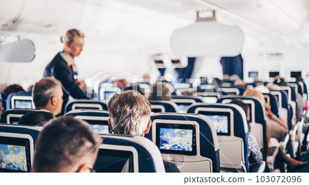 Interior of airplane with passengers on seats and stewardess in uniform walking the aisle, serving people. Commercial economy flight service concept 103072096