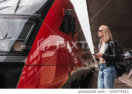 Young blond woman in jeans, shirt and leather jacket wearing bag and sunglass, embarking red modern speed train on train station platform. Travel and transportation 103074461