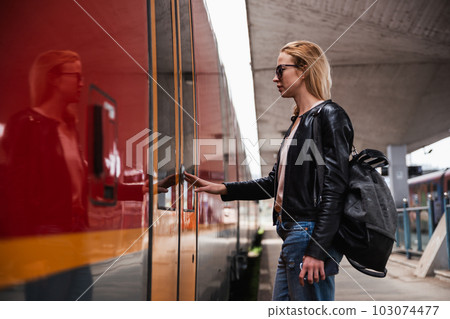 Young blond woman in jeans, shirt and leather jacket wearing bag and sunglass, presses door button of modern speed train to board on train station platform. Travel and transportation. 103074477