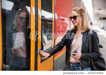 Young blond woman in jeans, shirt and leather jacket wearing bag and sunglass, presses door button of modern speed train to board on train station platform. Travel and transportation. Young blond woman in jeans, shirt and leather jacket wearing bag and sunglass, presses door button of modern speed train to board on train station platform. Travel and transportation. 103074482
