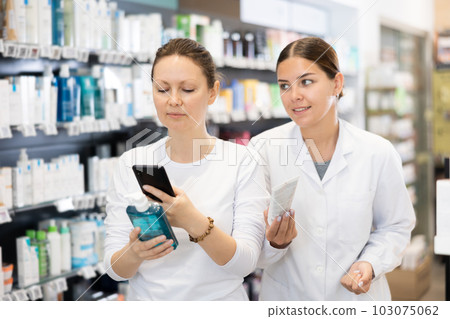 Woman scans QR code on packaging of face cream and listens attentively to consultant in pharmacy 103075062