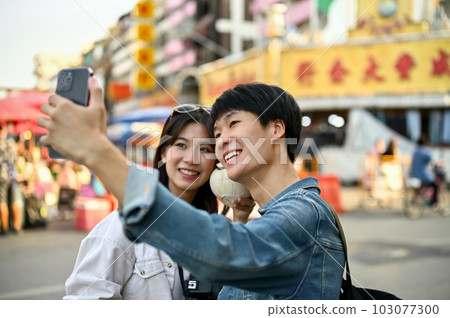 Happy young Asian tourist couple taking a photo with a smartphone in a Chinatown. 103077300