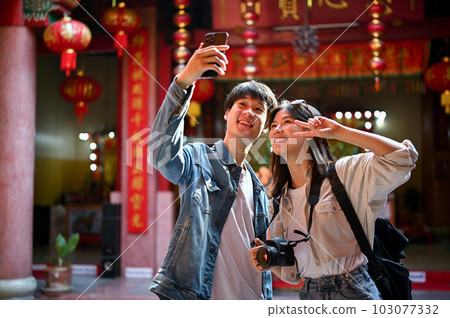 Cute Asian tourist couple taking their pictures with a smartphone while visiting a Chinese temple Cute Asian tourist couple taking their pictures with a smartphone while visiting a Chinese temple 103077332