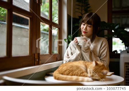 Relaxed young Asian woman sipping tea at a table with her orange cat. Relaxed young Asian woman sipping tea at a table with her orange cat. 103077745