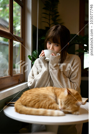 A woman in a cozy sweater resting with her orange cat and sipping tea in living room. 103077746