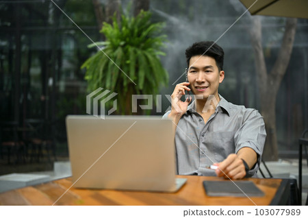 Smiling Asian businessman sitting outdoor and talking on the phone 103077989