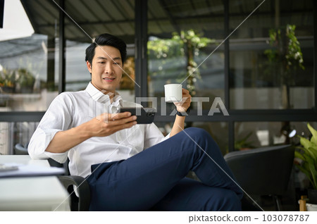 Relaxed Asian businessman watching videos on his phone while enjoying his coffee 103078787