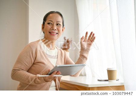 Attractive mature Asian woman sits in a coffee shop with her tablet. Attractive mature Asian woman sits in a coffee shop with her tablet. 103079447