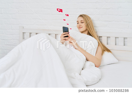 Young woman woke up and start browsing social media on mobile telephone while being in bed 103088286