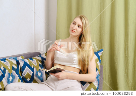 Pensive young woman takes notes in a notebook sitting at home on the couch 103088298