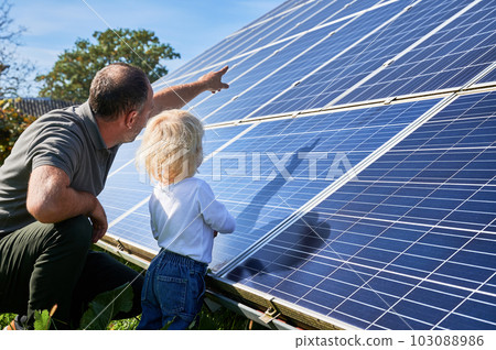 Man showing his small child the solar panels during sunny day. Father presenting to son modern energy resource. Back view of father and his kid watching on solar panels 103088986