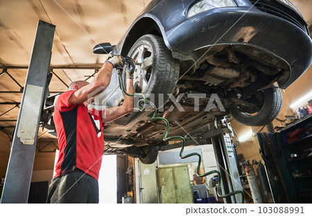 Mechanic, worker, repairman changing wheels of car in garage. Man wearing dirty gloves, working in servicing center, repairing cars. Concept of manual labor. Mechanic, worker, repairman changing wheels of car in garage. Man wearing dirty gloves, working in servicing center, repairing cars. Concept of manual labor. 103088991