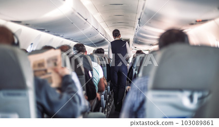 Interior of airplane with passengers on seats and stewardess in uniform walking the aisle, serving people. Commercial economy flight service concept 103089861