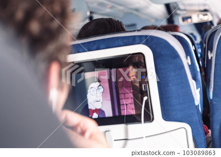 Reflection of a passenger on an airplane touch screen monitor while watching cartoon during long flight. Entertainment service system in aircraft Reflection of a passenger on an airplane touch screen monitor while watching cartoon during long flight. Entertainment service system in aircraft 103089863