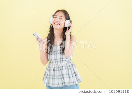 黃色背景下戴著耳機聽音樂時操作智能手機的年輕女性 黃色背景下戴著耳機聽音樂時操作智能手機的年輕女性 103091318
