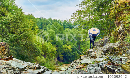 A young woman with an umbrella contemplates shallow mountain river in September 103092642