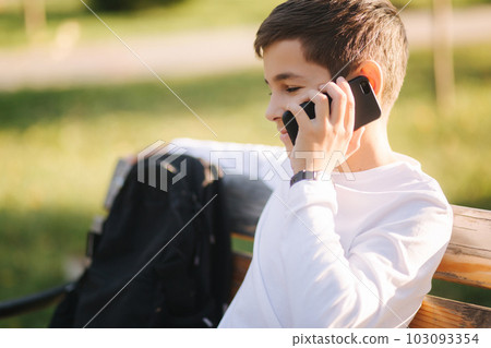 Young boy in white sweatshirt with black backpack sitting on the bench in the park and speak with somebody by the phone Young boy in white sweatshirt with black backpack sitting on the bench in the park and speak with somebody by the phone 103093354