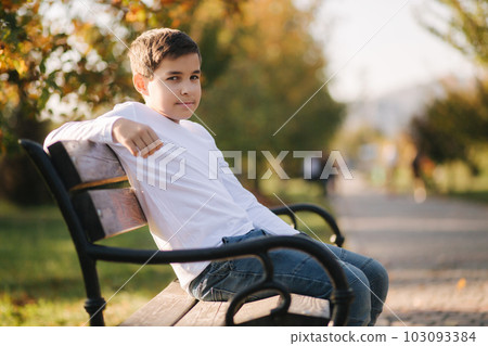 Teenage boy in white sweater sitting on the bench. Handsome school boy with backpack in autumn park Teenage boy in white sweater sitting on the bench. Handsome school boy with backpack in autumn park 103093384