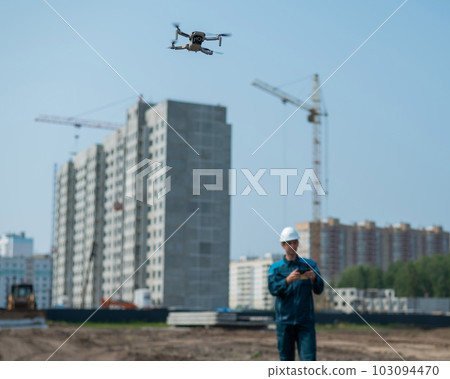 A man in a helmet and overalls controls a drone at a construction site. The builder carries out technical oversight. 103094470