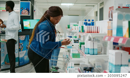 African american girl checking pharmaceutical products from pharmacy shelves, looking to buy medicaments and vitamins for healthcare. Young woman taking medicine and pills from drugstore. 103095247
