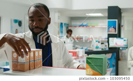 Pharmacy employee arranging stock of medicaments on drugstore shelves, putting boxes of pills and vitamins to help customers. Young man working with pharmaceutical products, retail store. 103095450