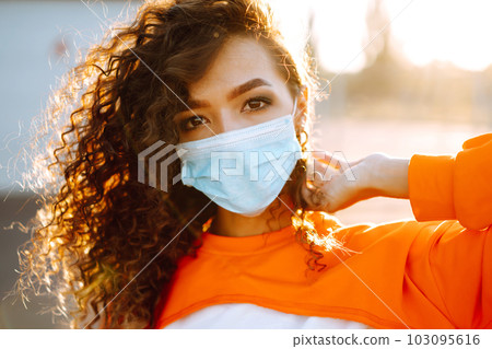 Young woman with medical face mask at sunset. Curly woman posing during quarantine of coronavirus pandemic. 103095616