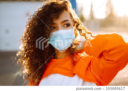 Young woman with medical face mask at sunset. Curly woman posing during quarantine of coronavirus pandemic. 103095617