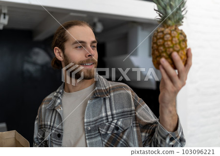 Bearded man in checkered shirt holding pineapples in hands standing in kitchen at home. Bearded man in checkered shirt holding pineapples in hands standing in kitchen at home. 103096221