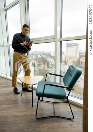 Dark-haired man with a phone in hands standing near the window Dark-haired man with a phone in hands standing near the window 103097043