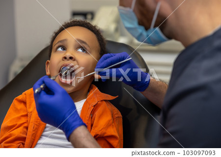 Dark-skinned boy looking scared while doctor checking up his teeth Dark-skinned boy looking scared while doctor checking up his teeth 103097093