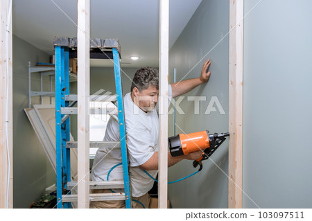 Construction worker uses an air hammer to nail wooden beams housing frames in preparation for construction. 103097511