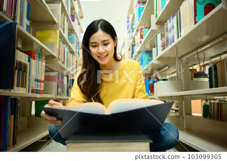 Education, learning, knowledge concept. Smiling female student sitting on floor in library between bookshelves, reading text book 103099305