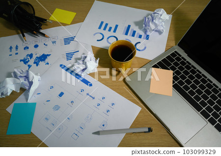 Cluttered office desk with crumpled papers, laptop, coffee cup and sticky notes 103099329