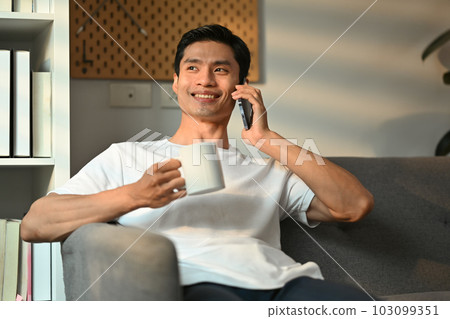 Positive millennial man having pleasant mobile phone conversation and drinking coffee in living room Positive millennial man having pleasant mobile phone conversation and drinking coffee in living room 103099351