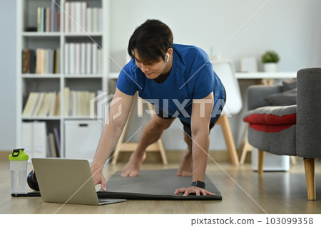Young man in sportswear working out at home and watching fitness lessons online on laptop. Fitness, training and healthy lifestyle Young man in sportswear working out at home and watching fitness lessons online on laptop. Fitness, training and healthy lifestyle 103099358