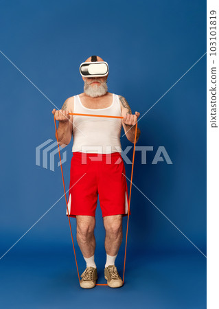 Portrait of mature, bearded man in sportswear and vr glasses training with fitness expanders against blue studio background Portrait of mature, bearded man in sportswear and vr glasses training with fitness expanders against blue studio background 103101819