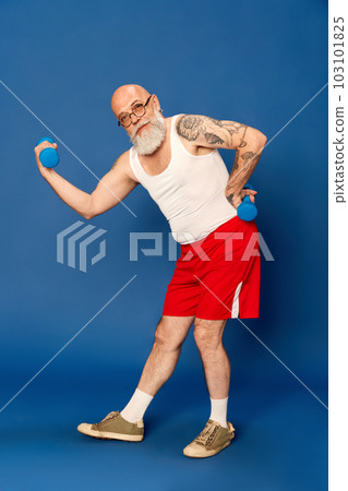 Portrait of mature, bearded man in sportswear training with dumbbells against blue studio background 103101825