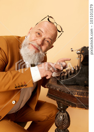 Portrait of bearded, mature, bald man in elegant, classical suit posing with typewriter against studio background. Happy novelist Portrait of bearded, mature, bald man in elegant, classical suit posing with typewriter against studio background. Happy novelist 103101879