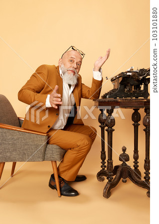 Portrait of bearded, mature, bald man in elegant, classical suit sitting near typewriter and spreading hands. Professional crisis 103101880