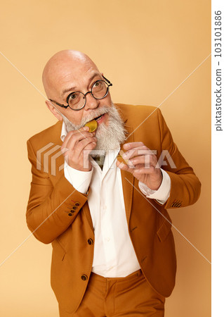 Portrait of bearded, mature, bald man in elegant, classical suit and glasses, biting gold coin, posing against studio background 103101886