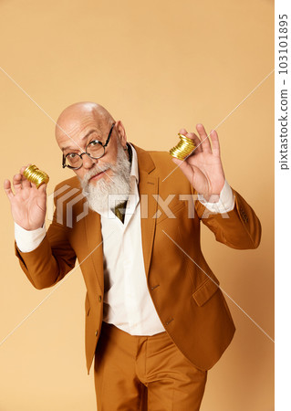 Portrait of bearded, mature, bald businessman in elegant, classical suit posing with golden coins against studio background Portrait of bearded, mature, bald businessman in elegant, classical suit posing with golden coins against studio background 103101895