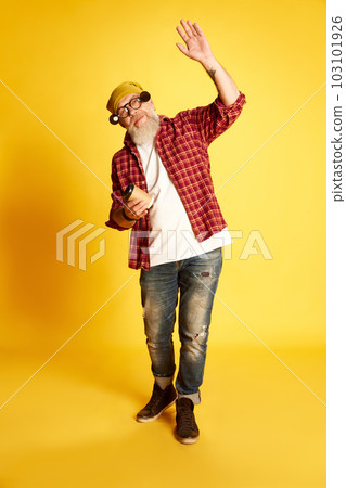 Full-length portrait of mature bearded man in stylish clothes, checkered shirt and hat, posing with coffee against yellow studio background 103101926