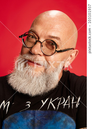 Portrait of bearded, mature, bald man in glasses and casual stylish clothes posing,m looking at camera and smiling against red studio background 103101937
