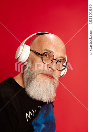Close-up portrait of mature, bearded, bald man in glasses, listening to music in headphones and looking at camera against red studio background 103101986