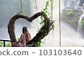 a young woman on a swing in the shape of a heart, enjoying the rainforest and looking at the high powerful waterfall Tegenungan. Rear view of a tourist girl on the Indonesian island of Bali 103103640
